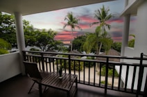 A serene view from a balcony overlooks a tropical landscape with palm trees and lush greenery. The sky is painted in vibrant hues of pink and purple, hinting at either sunrise or sunset. A wooden chair and table sit on the balcony, fostering a relaxing atmosphere, while thatched-roof structures are visible beyond the railing.