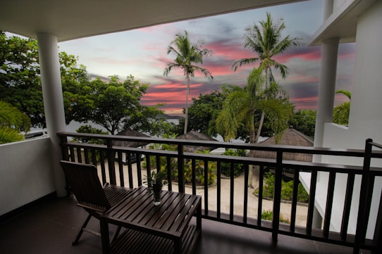 A serene view from a balcony overlooks a tropical landscape with palm trees and lush greenery. The sky is painted in vibrant hues of pink and purple, hinting at either sunrise or sunset. A wooden chair and table sit on the balcony, fostering a relaxing atmosphere, while thatched-roof structures are visible beyond the railing.
