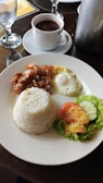 A plate of food featuring a serving of white rice, crispy fried pork belly, and a sunny-side-up egg, accompanied by a side salad of sliced tomato, cucumber, and lettuce on a white plate. A cup of hot coffee is placed on the table next to the plate, along with a glass of water.