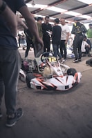 A group of people is gathered around a go-kart under a large metal canopy. The go-kart is in the foreground, predominantly white with red and black accents and labeled with the number 12. The individuals, some holding papers and wearing casual or racing attire, seem to be in discussion or preparation, possibly for a race event. The environment has a busy, bustling atmosphere.