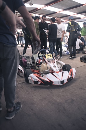 A group of people is gathered around a go-kart under a large metal canopy. The go-kart is in the foreground, predominantly white with red and black accents and labeled with the number 12. The individuals, some holding papers and wearing casual or racing attire, seem to be in discussion or preparation, possibly for a race event. The environment has a busy, bustling atmosphere.