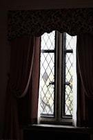 A vintage window with diamond-patterned glass panes framed by thick curtains. The curtains are dark and heavy, draping elegantly on either side. A floral valance runs across the top, adding an ornamental touch. Beyond the window, blurred greenery is visible, suggesting a garden or trees outside.