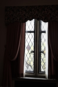 A vintage window with diamond-patterned glass panes framed by thick curtains. The curtains are dark and heavy, draping elegantly on either side. A floral valance runs across the top, adding an ornamental touch. Beyond the window, blurred greenery is visible, suggesting a garden or trees outside.