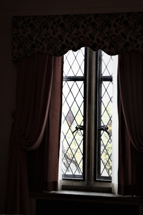 A vintage window with diamond-patterned glass panes framed by thick curtains. The curtains are dark and heavy, draping elegantly on either side. A floral valance runs across the top, adding an ornamental touch. Beyond the window, blurred greenery is visible, suggesting a garden or trees outside.