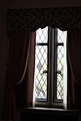 A vintage window with diamond-patterned glass panes framed by thick curtains. The curtains are dark and heavy, draping elegantly on either side. A floral valance runs across the top, adding an ornamental touch. Beyond the window, blurred greenery is visible, suggesting a garden or trees outside.