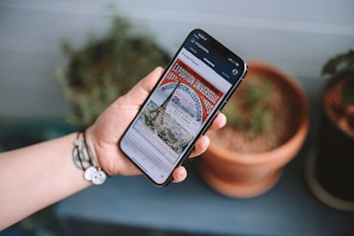 A hand with a bracelet holds a smartphone displaying an online article about the Exposition Universelle in Paris. The background features two terracotta pots with plants on a blue surface.