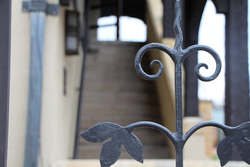 An ornamental iron fence is in focus, displaying intricate scrollwork and leaf motifs. In the background, a set of stairs leads up to a structure with beige walls and black architectural details. The image has a shallow depth of field, blurring the background.