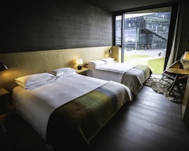 A modern hotel room featuring two neatly made beds with white linens and blankets in earthy tones. The room includes a minimalist wooden headboard and side tables with lamps. A large window provides a view of a modern building and grassy outdoor space, allowing natural light to fill the room. The interior is furnished with a dark wood floor, a desk with a chair, and a soft rug.