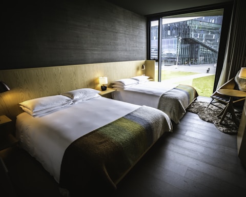 A modern hotel room featuring two neatly made beds with white linens and blankets in earthy tones. The room includes a minimalist wooden headboard and side tables with lamps. A large window provides a view of a modern building and grassy outdoor space, allowing natural light to fill the room. The interior is furnished with a dark wood floor, a desk with a chair, and a soft rug.