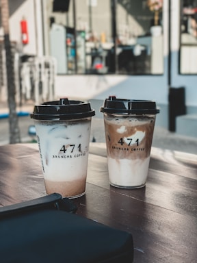 Two cups of coffee with lids are placed on a wooden table, featuring a modern and minimalist design from 471 Brunch & Coffee. The background includes blurred elements of a café setting, with large windows and reflections.