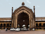 An elaborate and large historical arched gateway structure with intricate carvings and architecture. Several vehicles, including cars and motorcycles, are parked and moving in front of the structure. A few people can also be seen near the gateway. The sky in the background is clear with a few clouds.