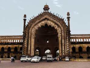 An elaborate and large historical arched gateway structure with intricate carvings and architecture. Several vehicles, including cars and motorcycles, are parked and moving in front of the structure. A few people can also be seen near the gateway. The sky in the background is clear with a few clouds.
