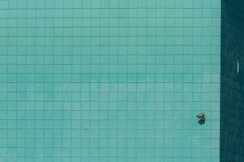 A grid of teal tiles covers the surface, with a single security camera mounted on the lower right. The tiles create a geometric pattern that is uniform across the wall.
