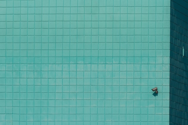 A grid of teal tiles covers the surface, with a single security camera mounted on the lower right. The tiles create a geometric pattern that is uniform across the wall.