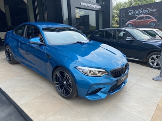 A shiny blue sports car is parked on a tiled floor in front of a dealership with a sign displaying 'roadway cars'. Another dark-colored SUV is parked nearby. The background features a billboard with a classic car image and the area is surrounded by greenery.