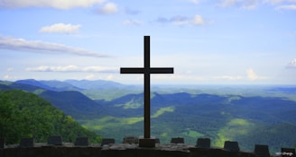 A large cross stands prominently at the edge of a stone platform overlooking a vast landscape of rolling hills and valleys. The sky is bright with scattered clouds, and the lush greenery of the hills extends into the distance.