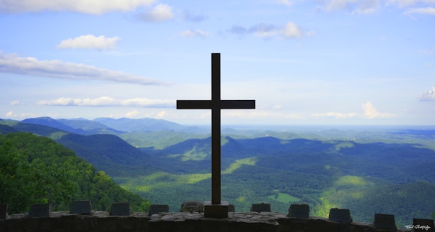 A large cross stands prominently at the edge of a stone platform overlooking a vast landscape of rolling hills and valleys. The sky is bright with scattered clouds, and the lush greenery of the hills extends into the distance.