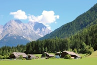 A picturesque village is nestled in a lush green valley, surrounded by dense forests and majestic mountains. Traditional houses with sloping roofs are scattered across the landscape. The sky is clear with a few puffy white clouds.