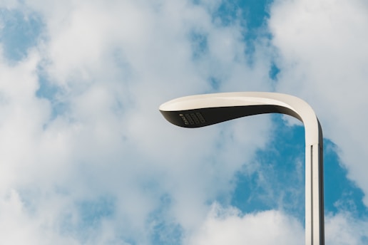 A modern streetlight stands against a backdrop of a partly cloudy sky. The streetlight has a sleek, curved design with a smooth finish, displaying simplicity and elegance.