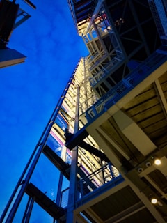A modern architectural structure with intricate metal beams and glass panels, viewed from a low angle against a striking blue sky. The design features a complex network of girders and supports, with reflective surfaces adding a dynamic interplay of light and shadow.