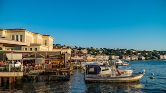 A picturesque waterfront scene with European-style architecture of light-colored buildings along the shore. Several boats are moored on the tranquil water. People are dining at open-air restaurants on the docks under canopies. The background is filled with lush greenery and additional buildings on rolling hills.