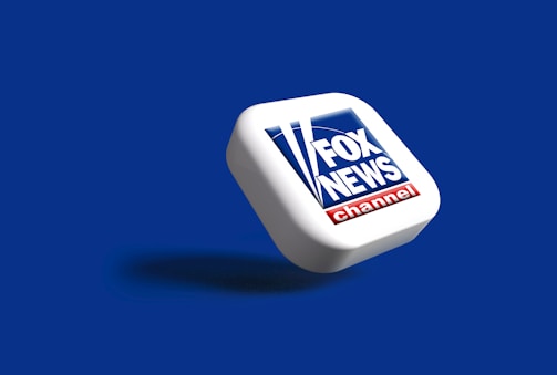 A three-dimensional representation of the Fox News Channel logo positioned diagonally on a blue background. The logo consists of the words 'FOX NEWS' in white text on a dark blue square, with red and white elements at the bottom.