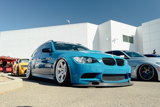 A low-angle view of several modified cars parked in a row at an outdoor event. The primary focus is on a bright blue BMW with a sporty, lowered stance and custom wheels. Behind it, there are other cars with similar modifications, including one in yellow and another in silver. The background consists of a large, white modern building and clear blue sky, with a few people visible in the distance.