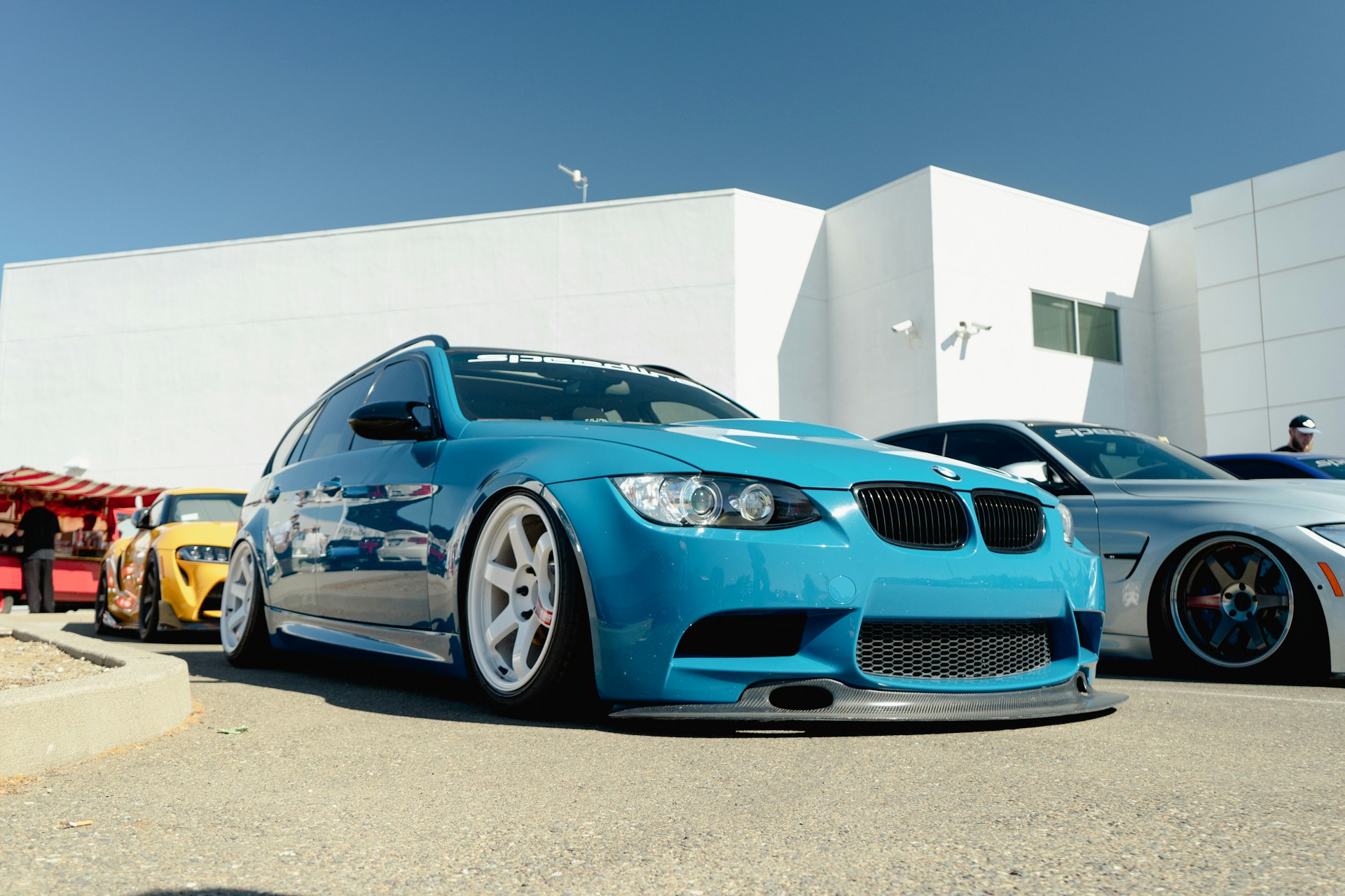 A low-angle view of several modified cars parked in a row at an outdoor event. The primary focus is on a bright blue BMW with a sporty, lowered stance and custom wheels. Behind it, there are other cars with similar modifications, including one in yellow and another in silver. The background consists of a large, white modern building and clear blue sky, with a few people visible in the distance.
