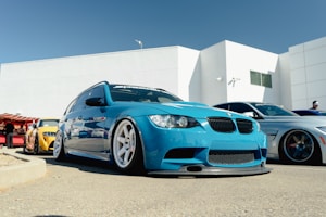 A low-angle view of several modified cars parked in a row at an outdoor event. The primary focus is on a bright blue BMW with a sporty, lowered stance and custom wheels. Behind it, there are other cars with similar modifications, including one in yellow and another in silver. The background consists of a large, white modern building and clear blue sky, with a few people visible in the distance.