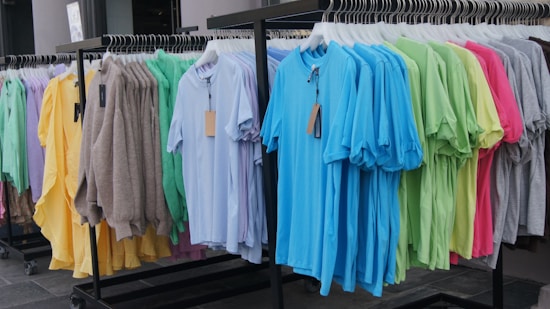 A clothing rack holds a variety of garments, including an assortment of colorful t-shirts and sweaters. The clothes are neatly hung on hangers, organized by color in vibrant shades such as blue, green, pink, yellow, and brown.