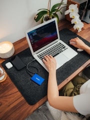 A person is working at a wooden desk with a laptop, using a wireless mouse. The desk also holds a smartphone, a portable hard drive, a candle, and a container with white orchids. A small speaker is positioned near the back.
