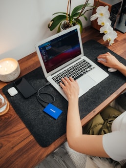A person is working at a wooden desk with a laptop, using a wireless mouse. The desk also holds a smartphone, a portable hard drive, a candle, and a container with white orchids. A small speaker is positioned near the back.
