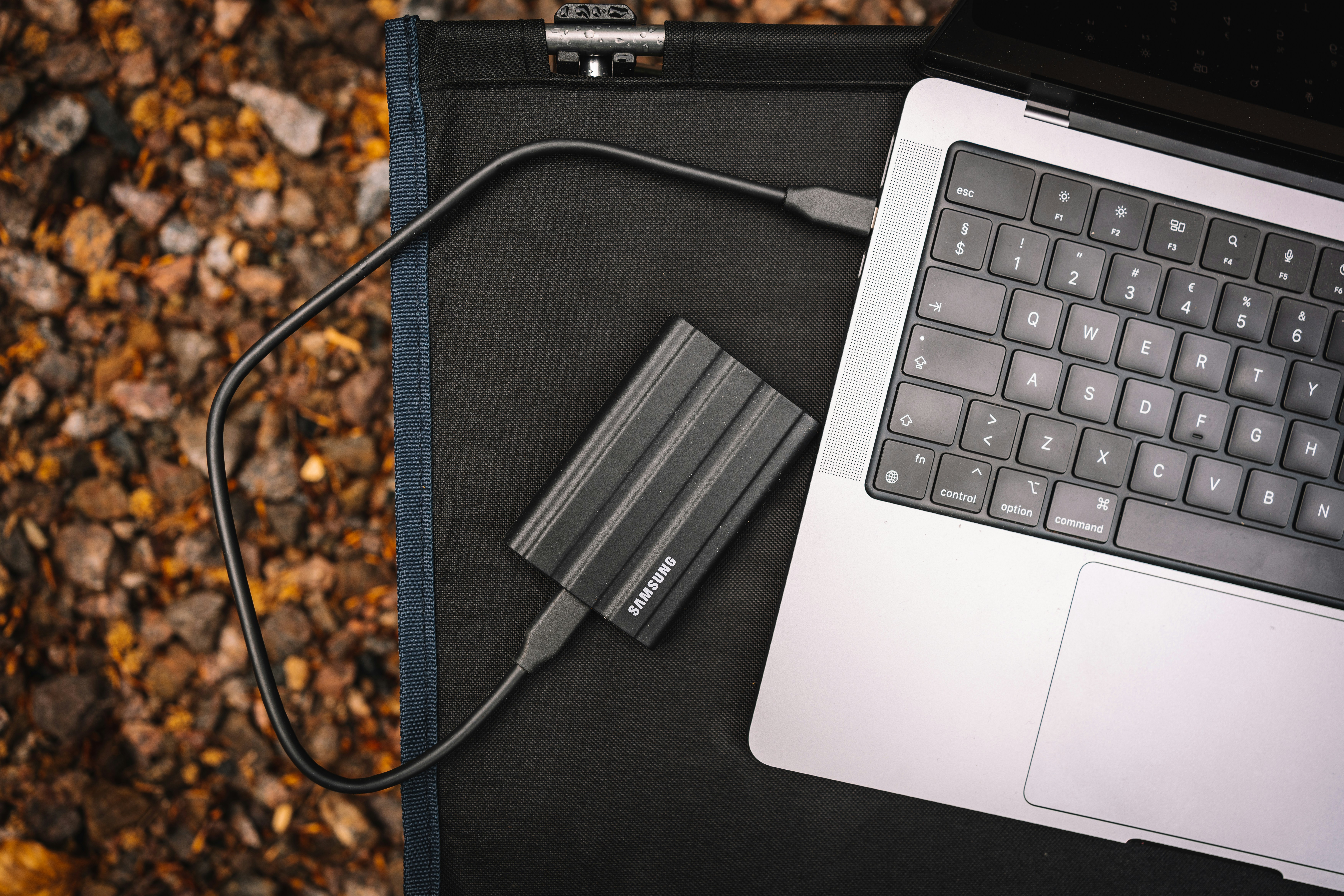 A portable Samsung SSD is connected by a black cable to a silver laptop on a dark mat.