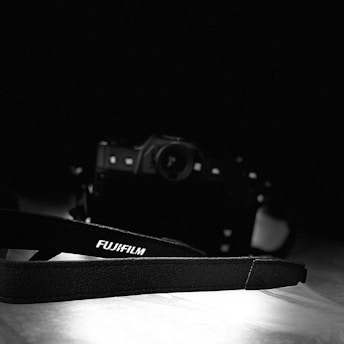 A black and white photograph displaying a Fujifilm camera in the background with a strap prominently featuring the Fujifilm logo in the foreground. The lighting highlights the texture of the strap, with the camera slightly out of focus, creating a moody ambiance.