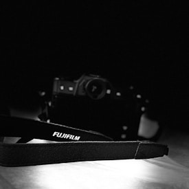 A black and white photograph displaying a Fujifilm camera in the background with a strap prominently featuring the Fujifilm logo in the foreground. The lighting highlights the texture of the strap, with the camera slightly out of focus, creating a moody ambiance.