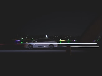 A car moves quickly along a dark road, with light trails emphasizing its speed. The background includes blurred lights from distant buildings or streetlights, suggesting nighttime or low light conditions.