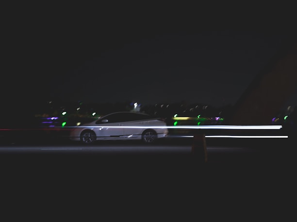A car moves quickly along a dark road, with light trails emphasizing its speed. The background includes blurred lights from distant buildings or streetlights, suggesting nighttime or low light conditions.