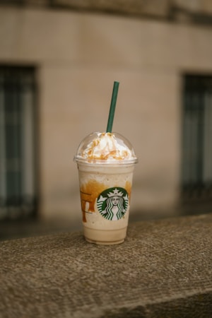 A Starbucks frappuccino cup is placed on a stone ledge. The cup is filled with a creamy, caramel-colored beverage and topped with whipped cream and caramel drizzle. A green straw protrudes from the dome lid of the cup.