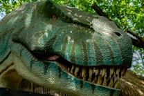 A close-up of a realistic dinosaur model with intricate scales and sharp teeth, positioned against a backdrop of lush green foliage. The sunlight filters through the leaves, casting dappled light shadows on the dinosaur’s textured skin.
