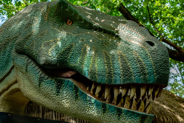 A close-up of a realistic dinosaur model with intricate scales and sharp teeth, positioned against a backdrop of lush green foliage. The sunlight filters through the leaves, casting dappled light shadows on the dinosaur’s textured skin.