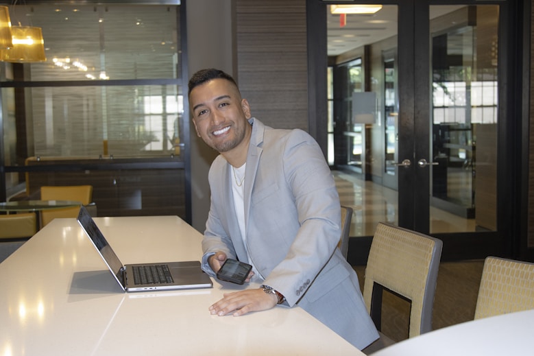 A person in a light gray suit leans on a table while smiling at the camera. The setting appears to be a modern office or conference area with large windows, glass doors, and soft lighting. A laptop is open on the table, and the person is holding a smartphone in their hand.