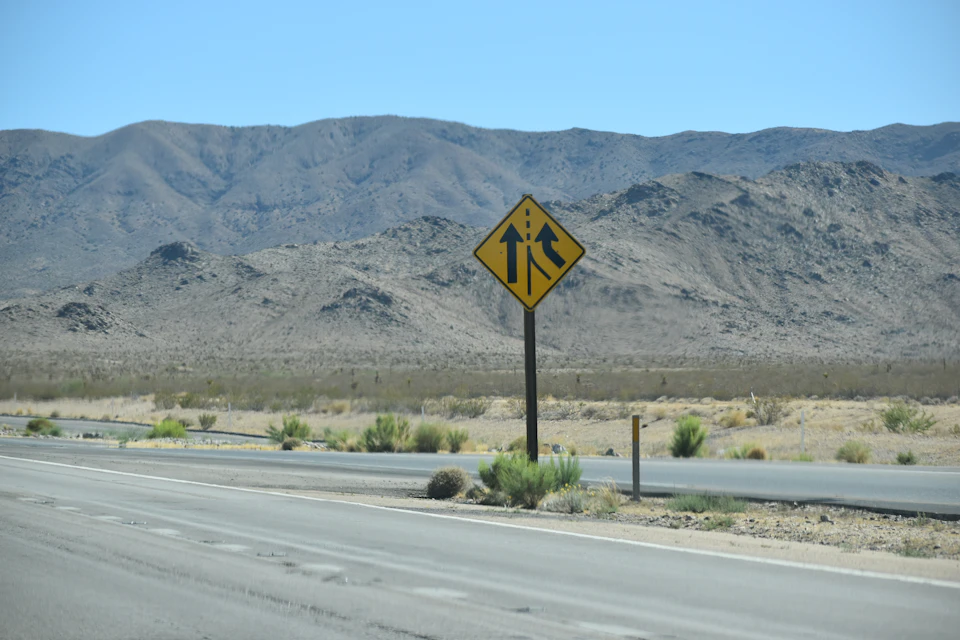 The image shows a desert road with a yellow diamond-shaped sign mounted on a wooden pole, displaying two black arrows pointing to the right and a warning for pedestrians. The road is surrounded by mountains under a clear blue sky, creating a striking contrast between the natural landscape and human-made infrastructure.