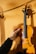 A hand is holding a violin against a textured surface, likely a corkboard. The violin has a glossy, varnished wood finish and a black fingerboard. The lighting creates shadows and highlights, emphasizing the contours and wood grain of the instrument.