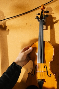 A hand is holding a violin against a textured surface, likely a corkboard. The violin has a glossy, varnished wood finish and a black fingerboard. The lighting creates shadows and highlights, emphasizing the contours and wood grain of the instrument.