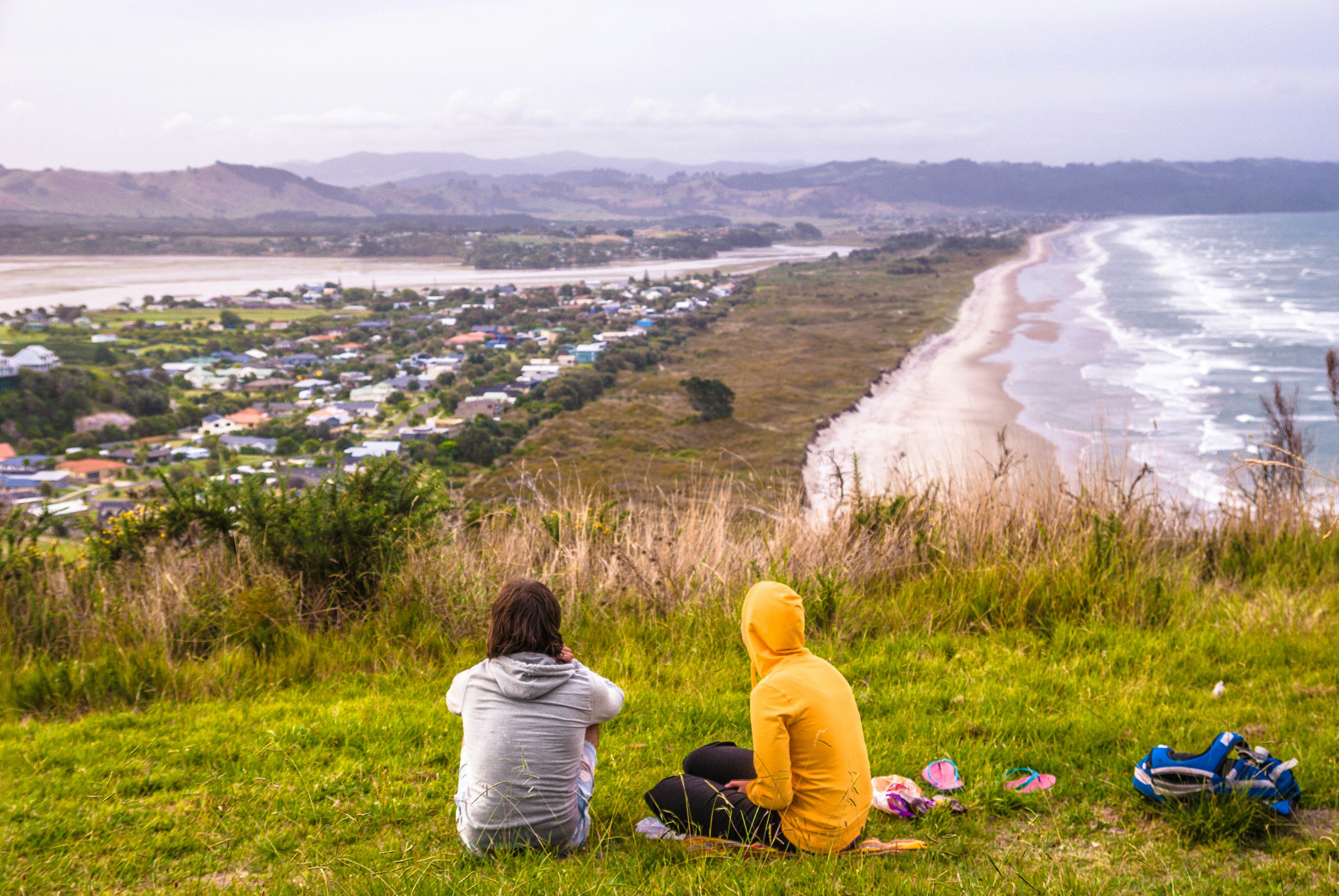 two people sitting on a hill overlooking a beach and town