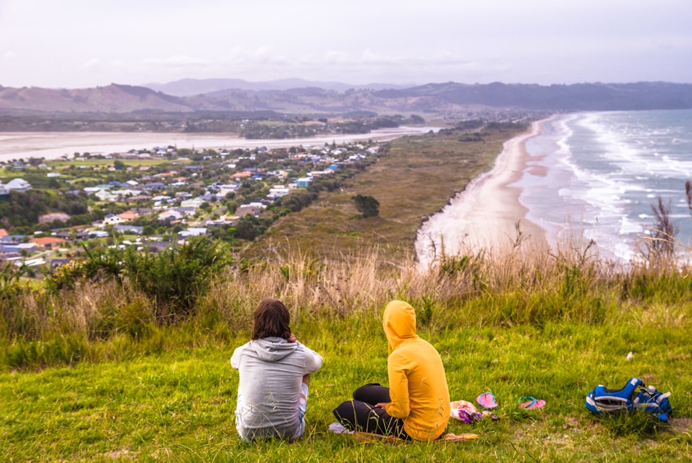 two people sitting on a hill overlooking a beach and town
