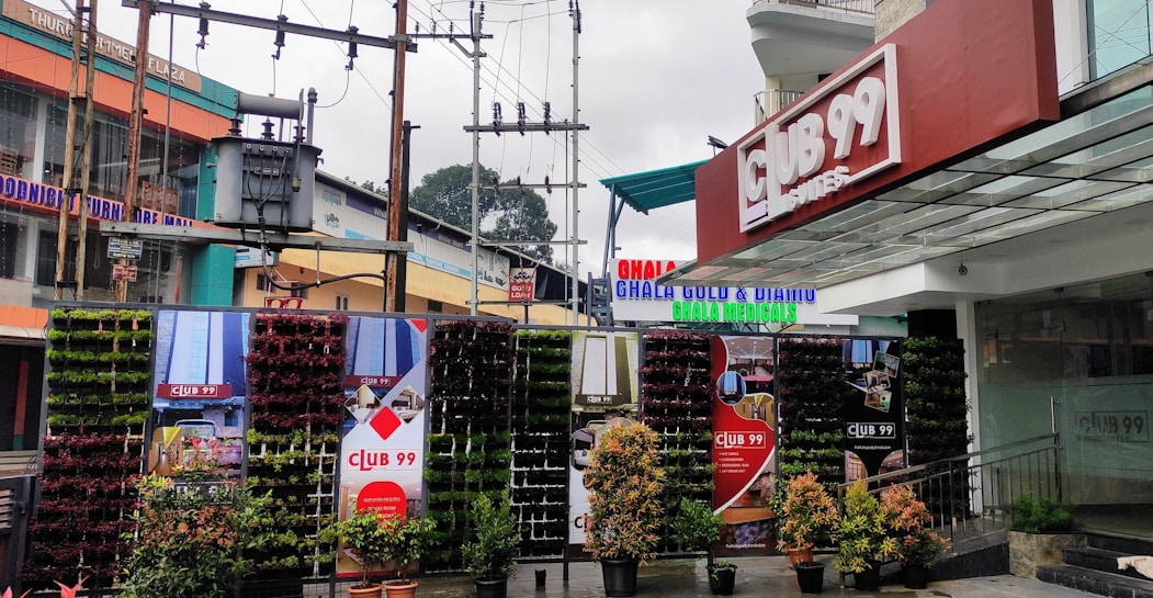 A storefront with the sign 'Club 99' in a modern commercial area. Vertical plant walls with various green and reddish plant pots are arranged at the entrance. Above, there are electrical transformers and cables. Surrounding buildings include shops with colorful signs, including one for 'Ghala Gold & Diamond'. The setting is urban, with overcast skies suggesting a cool or rainy atmosphere.