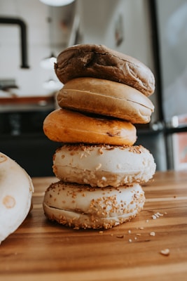 A stack of five bagels in various colors, topped with seeds, sits neatly arranged on a wooden surface. The bagels are of different shades, ranging from light beige to dark brown, and they are placed in a well-lit indoor environment with soft focus on the background.