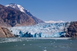 A vast, striking glacier meets the sea, with rugged mountains rising on either side. The ice is a bright, crystalline blue, contrasting with the dark, rocky surfaces of the mountains. Snow-capped peaks are visible in the background, under a clear blue sky.
