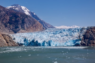 A vast, striking glacier meets the sea, with rugged mountains rising on either side. The ice is a bright, crystalline blue, contrasting with the dark, rocky surfaces of the mountains. Snow-capped peaks are visible in the background, under a clear blue sky.